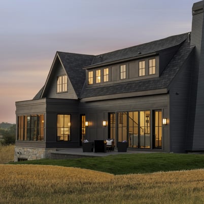 A gray brick farmhouse sits at sundown, with large black windows reflecting indoor light. 